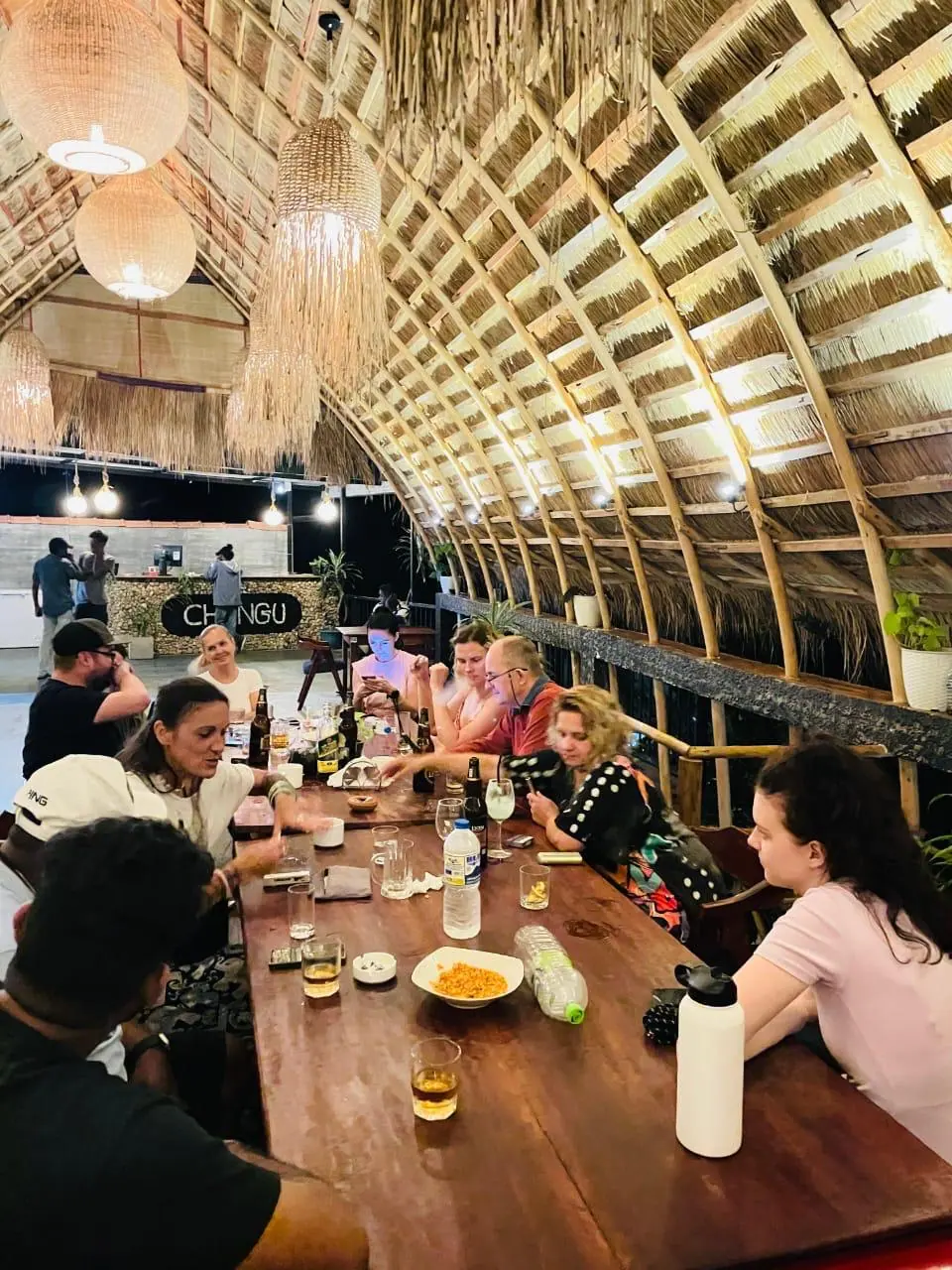 Guests dining at a long wooden table beneath Chingu's thatched roof and woven lamps